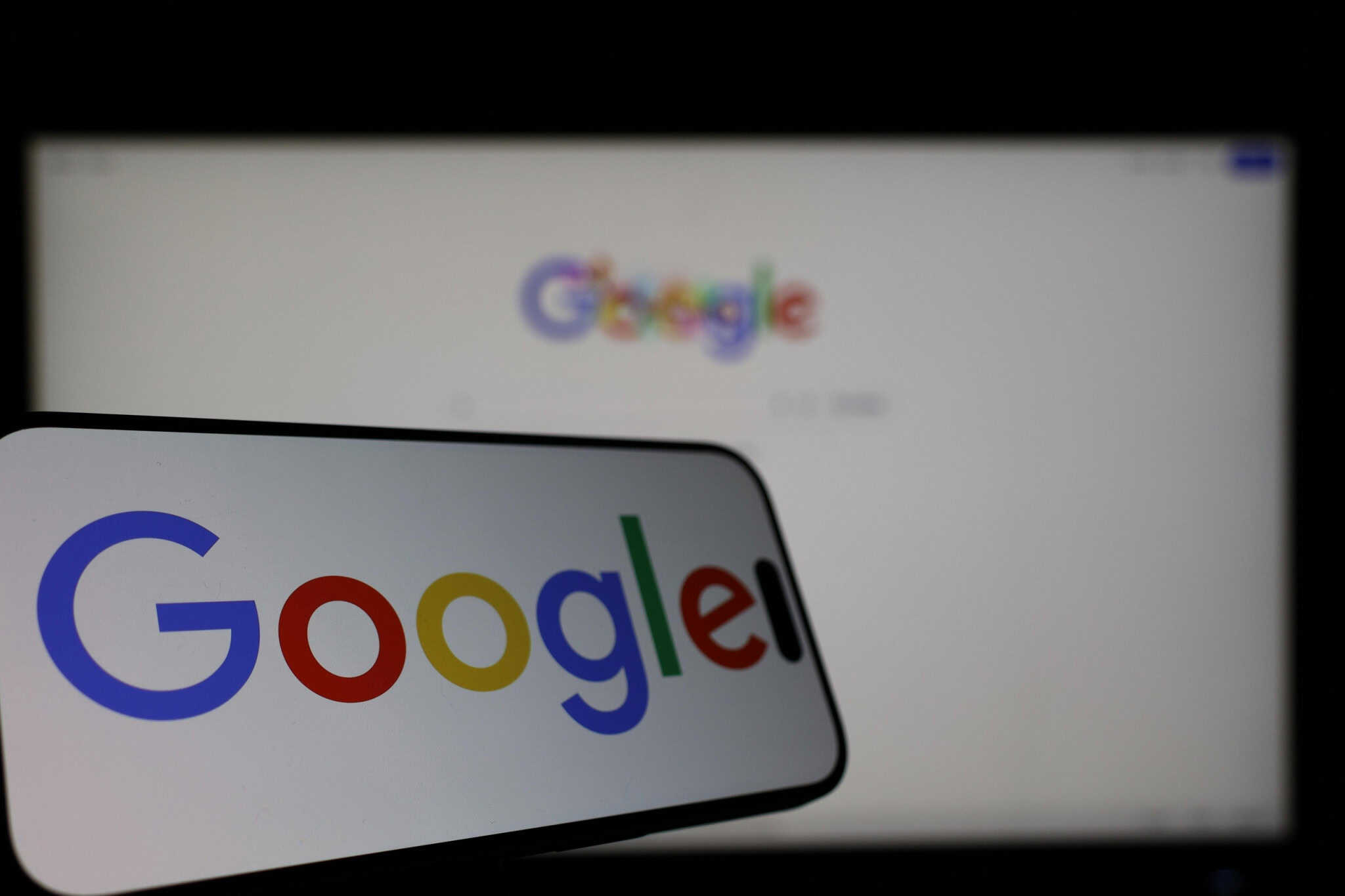 Ao fundo, desfocado, página do Google na internet; à frente, logo do google em um smartphone posicionado na horizontal