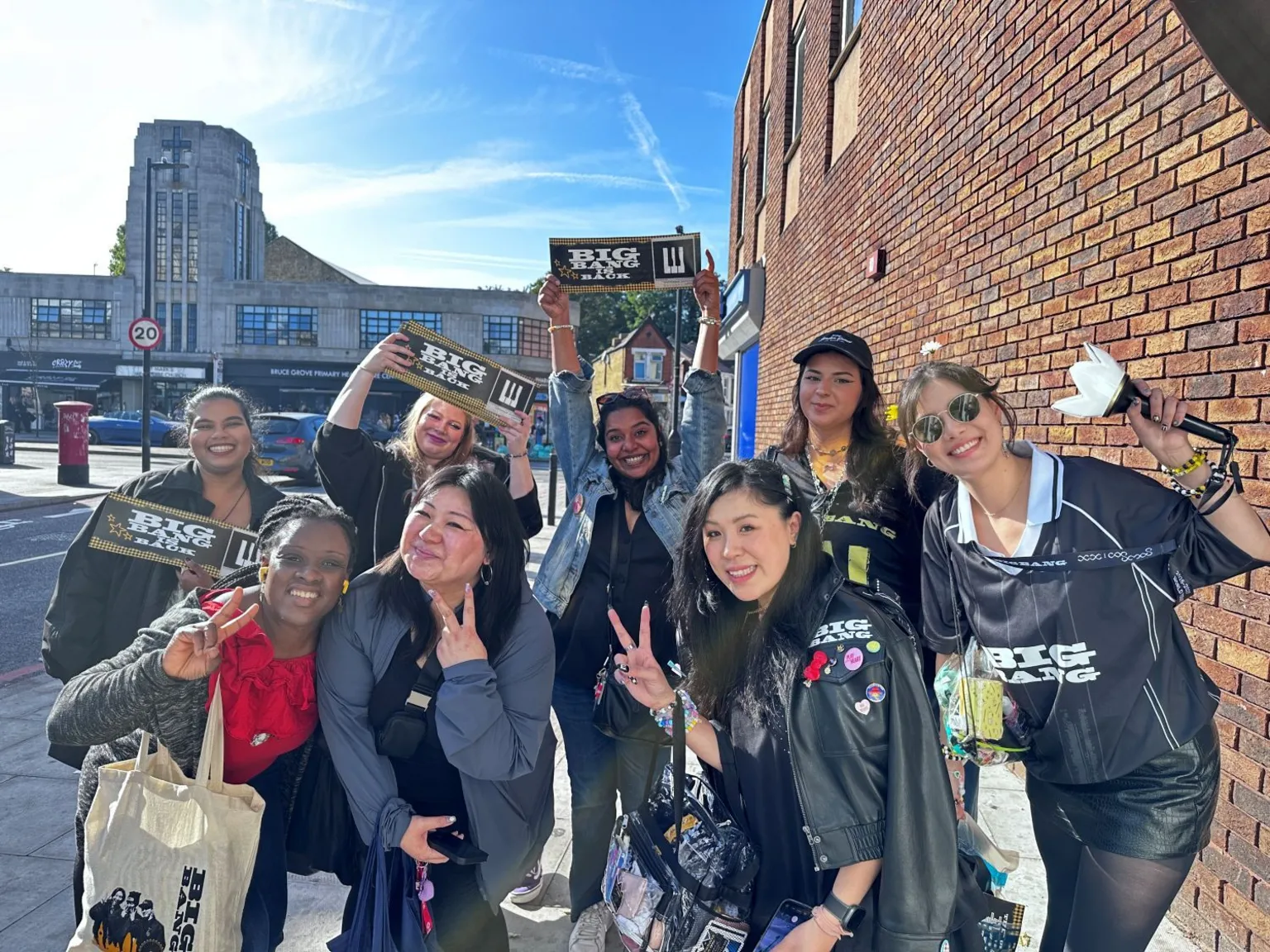Eight women gathered on a pavement beside a brick wall pose with concert merchandise. Several hold rectangular banners reading “BIG BANG IS BACK”, while one person holds a white flower-shaped light stick. Some clothing displays BIGBANG branding, and several accessories and badges are attached to bags.