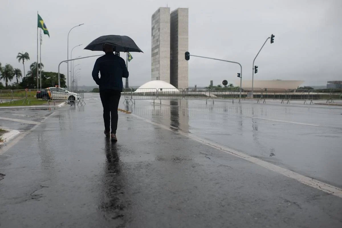 Imagem colorida de um homem caminhando em meio à chuva, usando guarda-chuva. Ao fundo, o Congresso Nacional