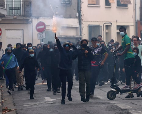 Masked students march in a street protest, one holding up a lit flare with smoke