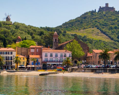 A small town of pastel buildings alongside a sandy shore and backed with green hills with a fort and a windmill on them