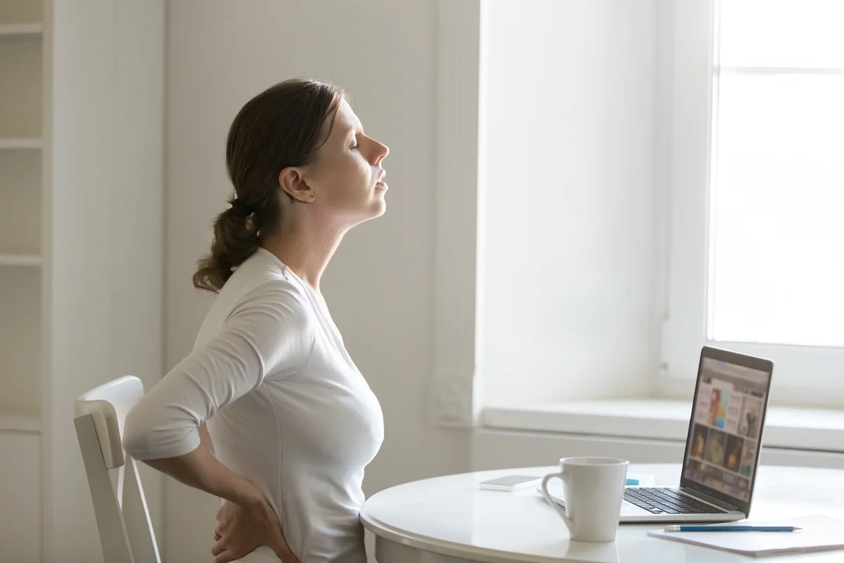 Imagem mostra mulher de roupas claras sentada em uma mesa e alongando as costas para melhorar a postura- Metrópoles